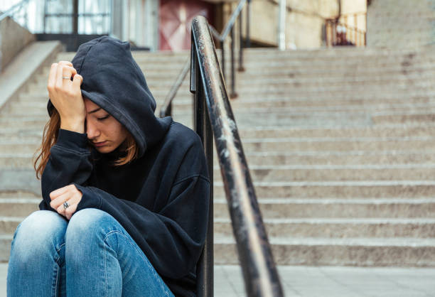 Depressed young woman sitting on the street Teenage girl sitting outdoors on the staircase covering her head with sweatshirt hood sulking stock pictures, royalty-free photos & images