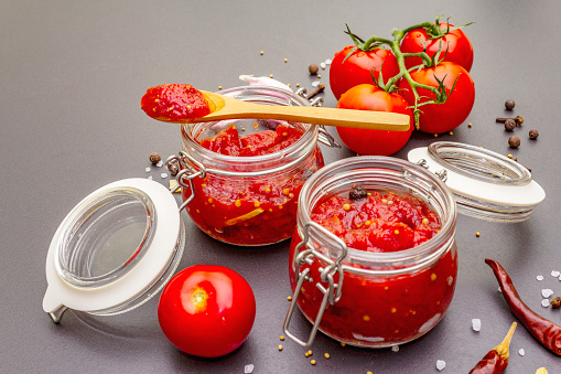 Tomato confiture, jam, chutney, sauce in glass jar. Homemade preservation concept. Fresh tomatoes, dried chili, spices, mustard beans, sea salt. On a stone background