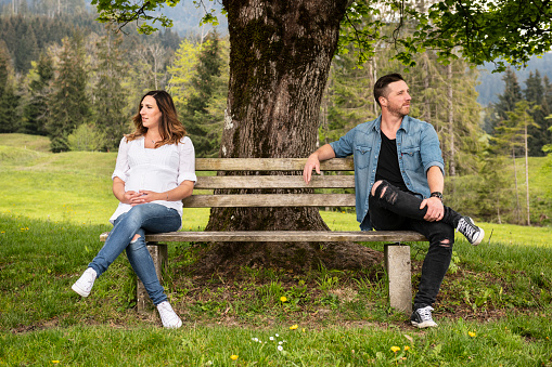 Pregnant woman sitting on one side of a bench and her boyfriend on the other side. They are looking in different directions. Concept: dispute, separation