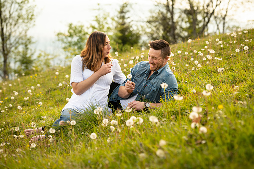 Pregnant woman sitting with her husband in a meadow. They are having fun with some dandelions.