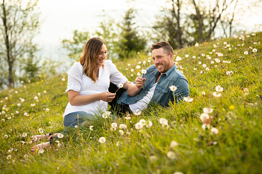 Pregnant woman sitting with her husband in a meadow. They are having fun with some dandelions.