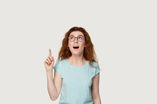 Astonished redhead woman pointing finger up at copyspace studio shot