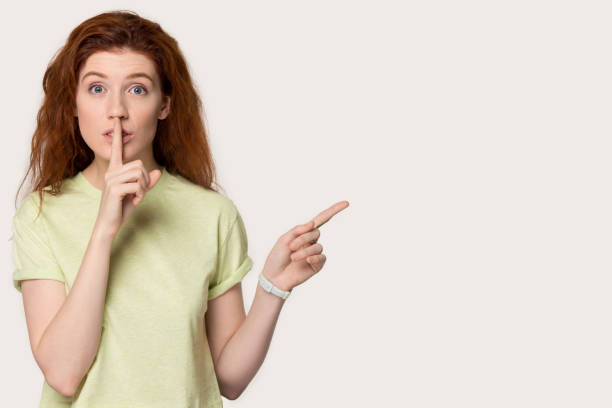 istock Redhead woman making hush gesture pointing finger aside at copyspace 1167561664