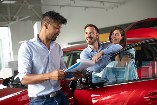 Husband and wife talking to a car salesman about buying a new car in a dealership showroom. Husband is leaning on a car door, while his wife is by his side, her hands on his shoulder. Car salesman has a catalog in his hands.