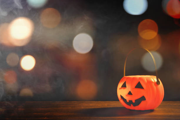 istock Halloween concept - Orange plastic pumpkin lantern on a dark wooden table with blurry sparkling light in the background, trick or treat. 1167576296