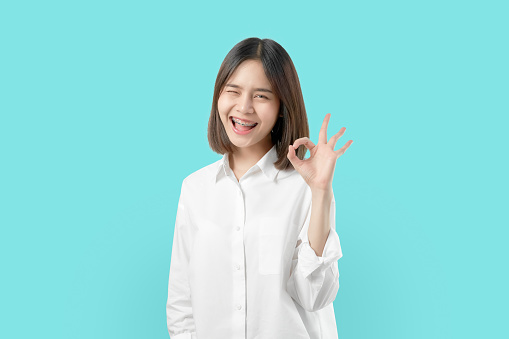 Portrait happily Asian woman shows ok sign and braces smiling witch looking at the camera on blue background, Cheerful girl gestures studio.