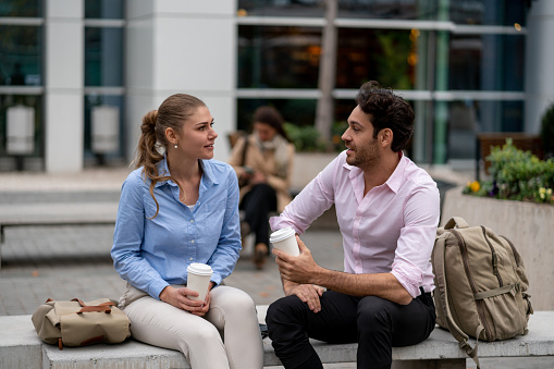 Young business couple outside enjoying a coffee sitting on bench talking - Focus on foreground