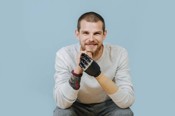 Young Man With Artificial hand is looking forward and smiling at the camera Portrait Of a smiling Man With Artificial hand in casual clothes sitting at the chair and looking at the camera. Studio shot. Isolated blue background prosthetic equipment photos stock pictures, royalty-free photos & images