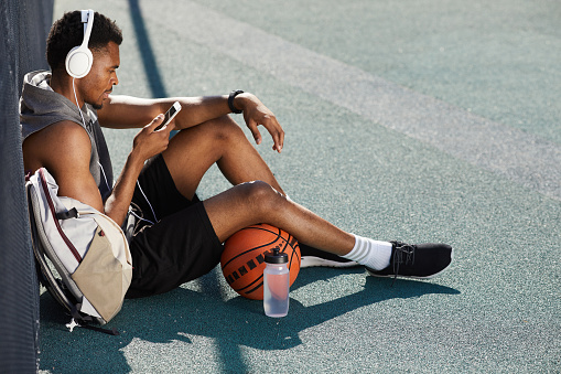 High angle portrait of contemporary African-American man wearing headphones and using smartphone while sitting in basketball court outdoors, copy space