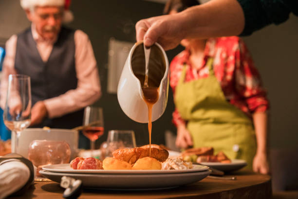 Completing The Meal With Gravy Gravy being poured from a jug onto a plate of Christmas lunch. A woman is wearing an apron out of focus in the background and there are more plates and glasses on the table ready for the family to sit down. gravy photos stock pictures, royalty-free photos & images