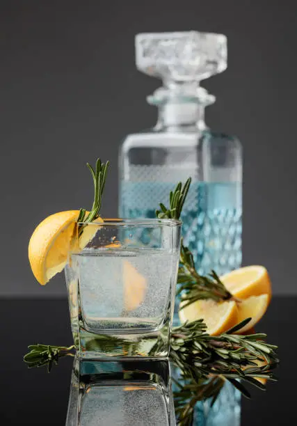 Cocktail gin-tonic with lemon slices and twigs of rosemary on a black reflective background.