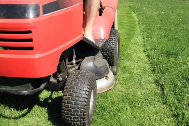 Red riding lawn mower (lawnmower, ride-on mower) in garden. Part of grass is mowed, piece is high with flowers. Concept close up wheels and cutting deck (mowing deck) when seasonal gardening.