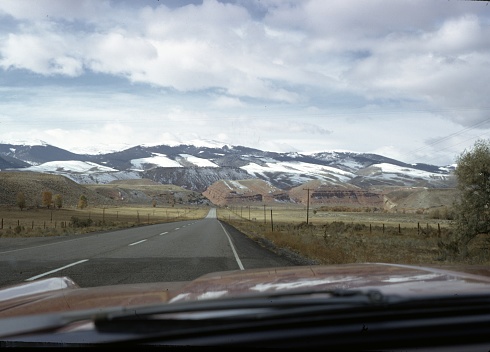 Photograph of a highway in a rural area with a mountain range partly covered with snow, on a cloudy day in Wyoming, 1970