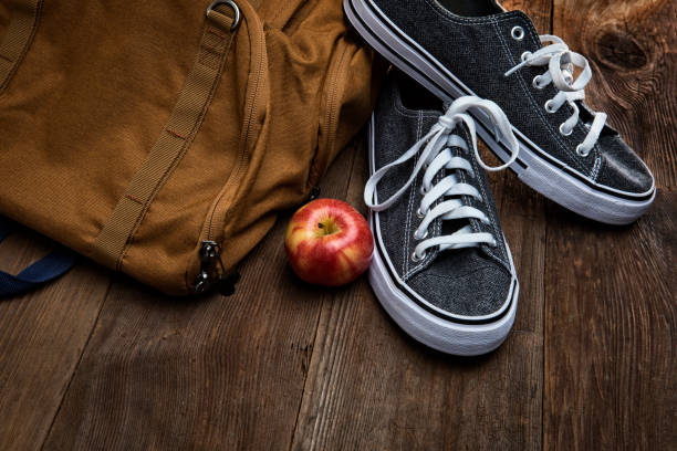 This is an overhead photograph of a backpack on a wood canvas of retro planks. It is surrounded by sneakers and a school supplies