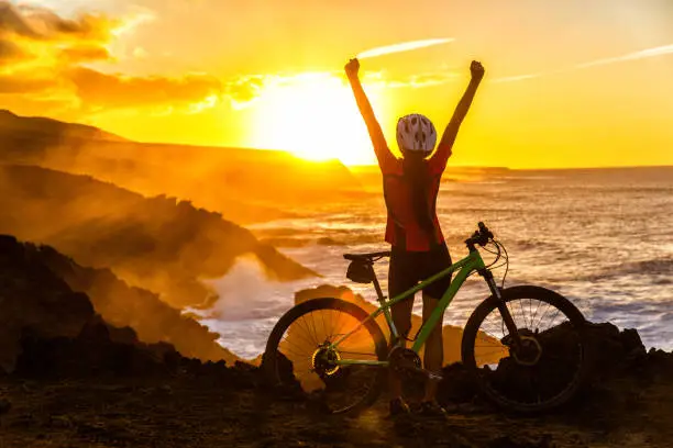 Success, achievement, accomplishment and winning concept with cyclist mountain biking. Winning happy MTB woman cycling reaching goal raising arms at sunset cheering and celebrating at summit top.