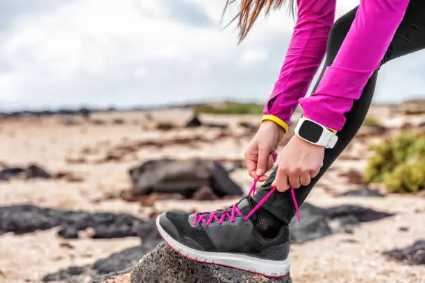 Fitness smartwatch woman runner lacing running shoes on beach, Athlete girl getting ready for run workout tying running shoe laces outside wearing watch gear. Healthy lifestyle concept.