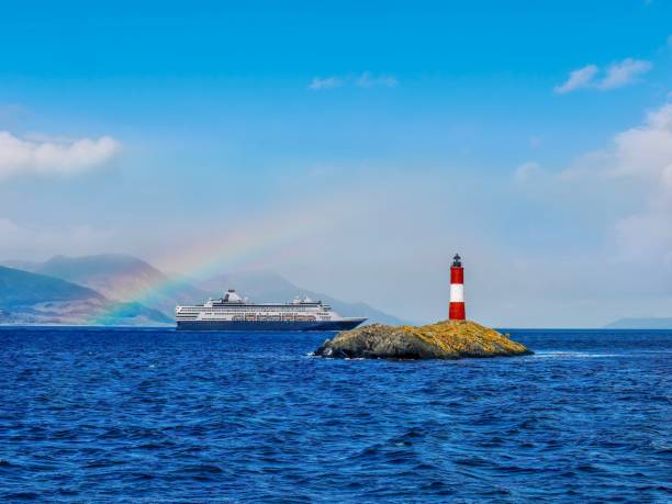 A small island in the Beagle Channel near Ushuaia, Argentina, with a popular tourist attraction of Les Eclaireurs (The Scouts) Lighthouse. A cruise ship and rainbow are in the background. A picturesque scene, with a red and white lighthouse on a small rocky island in a tranquil, blue channel. A cruise ship in the background is framed by a partial rainbow, which is set against a vibrant blue sky with some fluffy clouds. This area is the gateway to Antarctica from southern Argentina. beagle channel stock pictures, royalty-free photos & images