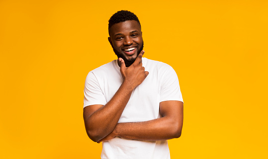 Interested african american man touching chin and smiling, yellow studio background, copy space