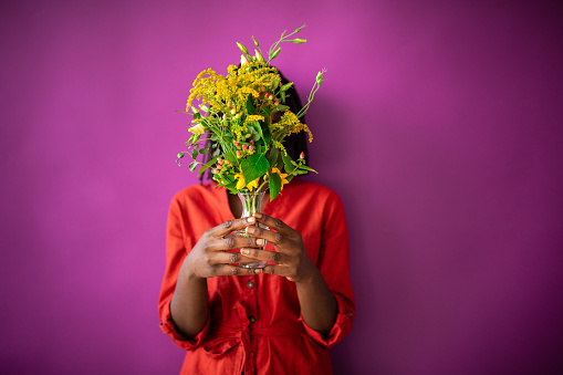 Unrecognizable woman standing in front of purple wall and holding bouquet of flowers in vase in her hands