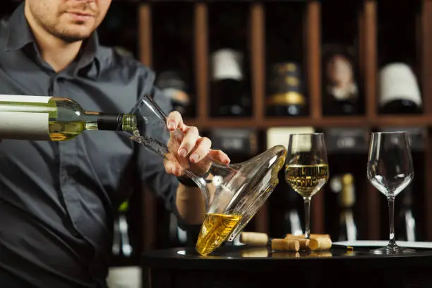 Close up portrait of caucasian sommelier pouring white wine from opened bottle in decanter designed to degustate and taste alcohol beverages, cellar full with containers collection having labels placed on shelves
