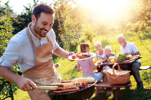 Food, people and family time concept.Young man grilling sausages on barbecue at summer garden party .