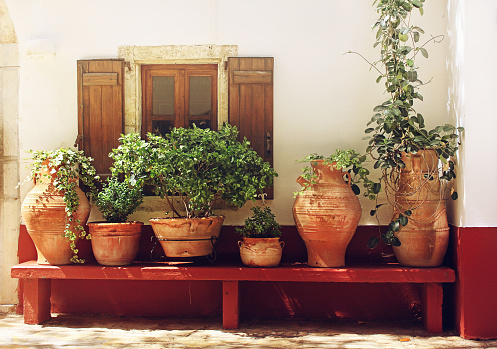 Picturesque textured wall with a window and plants in ceramic flower pots on a bench, sunlit, detail of small church in Greece, Crete