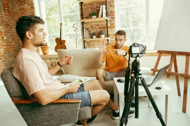 Photo of Two young male bloggers with professional camera recording video interview at home