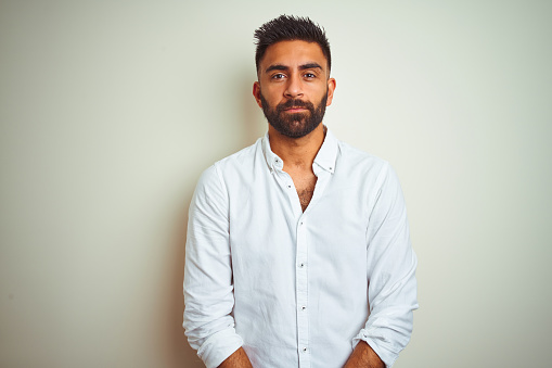 Young indian man wearing elegant shirt standing over isolated white background with serious expression on face. Simple and natural looking at the camera.