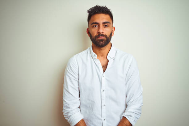 young indian man wearing elegant shirt standing over isolated white background with serious expression on face. simple and natural looking at the camera. - asian and indian ethnicities imagens e fotografias de stock