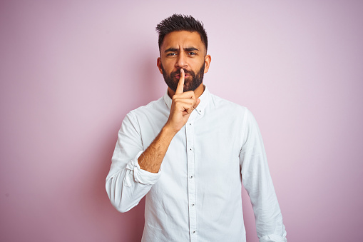 Young indian businessman wearing elegant shirt standing over isolated pink background asking to be quiet with finger on lips. Silence and secret concept.