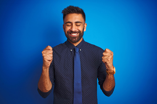 Young indian businessman wearing elegant shirt and tie standing over isolated blue background excited for success with arms raised and eyes closed celebrating victory smiling. Winner concept.