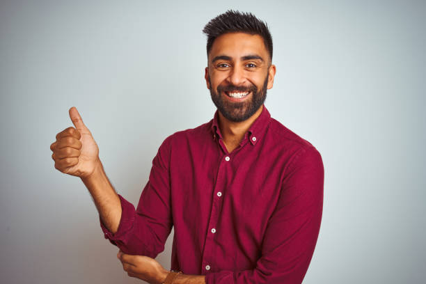 istock Young indian man wearing red elegant shirt standing over isolated grey background Looking proud, smiling doing thumbs up gesture to the side 1167769078