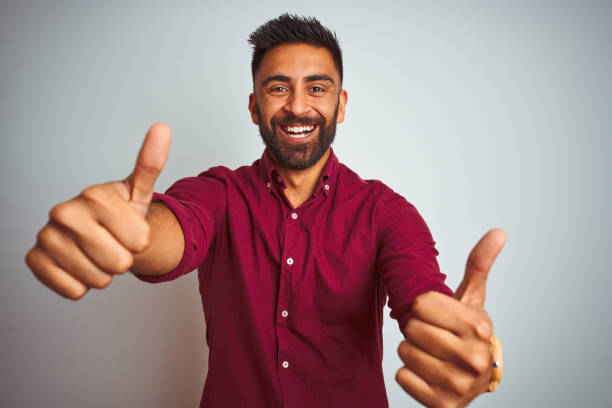 istock Young indian man wearing red elegant shirt standing over isolated grey background approving doing positive gesture with hand, thumbs up smiling and happy for success. Winner gesture. 1167769089