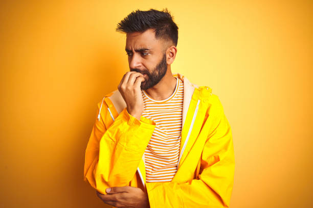 istock Young indian man wearing raincoat standing over isolated yellow background looking stressed and nervous with hands on mouth biting nails. Anxiety problem. 1167770156
