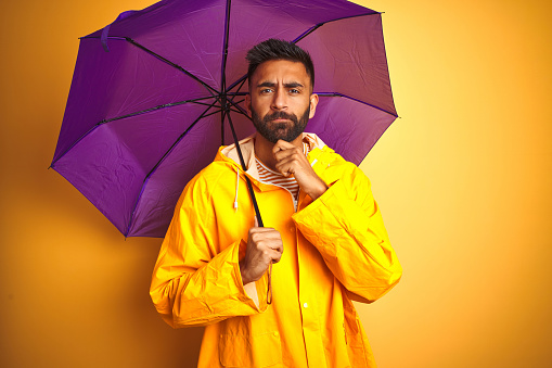 Young indian man wearing raincoat and purple umbrella over isolated yellow background serious face thinking about question, very confused idea