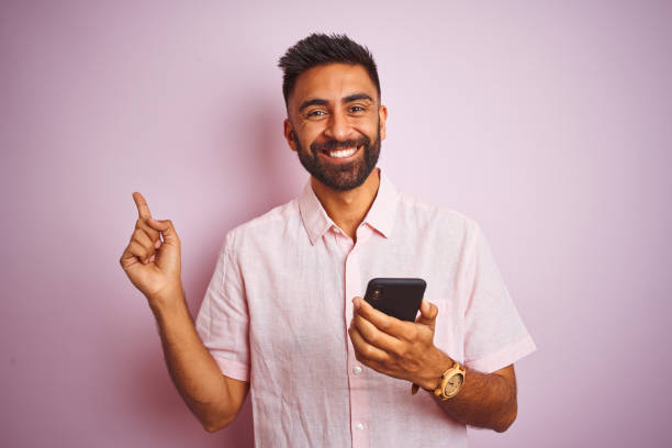 istock Young indian man using smartphone standing over isolated pink background very happy pointing with hand and finger to the side 1167770236