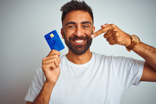 Young indian customer man holding credit card standing over isolated white background very happy pointing with hand and finger