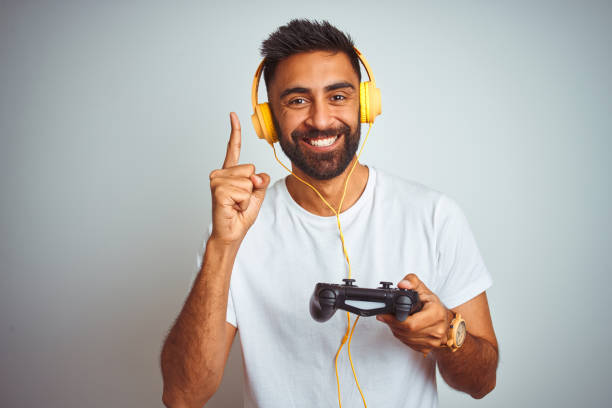 Arab indian gamer man playing video game using headphones over isolated white background surprised with an idea or question pointing finger with happy face, number one