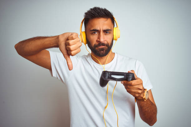 istock Arab indian gamer man playing video game using headphones over isolated white background with angry face, negative sign showing dislike with thumbs down, rejection concept 1167770700