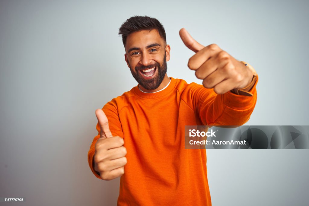 Jeune homme indien utilisant le chandail orange au-dessus du fond blanc d'isolement approuvant faisant le geste positif avec la main, pouces vers le haut souriant et heureux pour le succès. Geste du gagnant. - Photo de Bonheur libre de droits