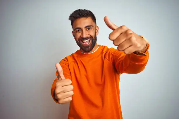 Photo of Young indian man wearing orange sweater over isolated white background approving doing positive gesture with hand, thumbs up smiling and happy for success. Winner gesture.