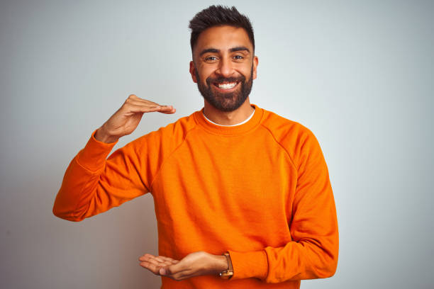 istock Young indian man wearing orange sweater over isolated white background gesturing with hands showing big and large size sign, measure symbol. Smiling looking at the camera. Measuring concept. 1167770711