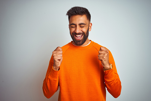 Young indian man wearing orange sweater over isolated white background excited for success with arms raised and eyes closed celebrating victory smiling. Winner concept.