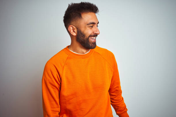 junger indischer mann trägt orangefarbenen pullover über isoliertem weißen hintergrund und schaut mit lächeln auf dem gesicht, natürlichem ausdruck zur seite. lachen zuversichtlich. - smiling single object photography orange stock-fotos und bilder