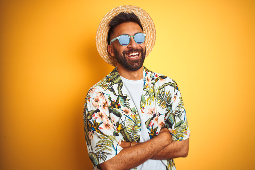 Hombre indio en vacaciones con gafas de sol de sombrero de camisa floral sobre fondo amarillo aislado cara feliz sonriendo con los brazos cruzados mirando a la cámara. Persona positiva. photo