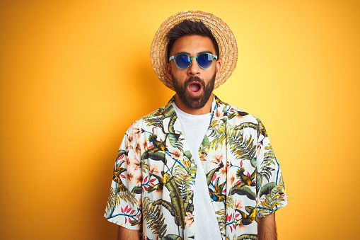 Indian man on vacation wearing floral shirt hat sunglasses over isolated yellow background afraid and shocked with surprise expression, fear and excited face.