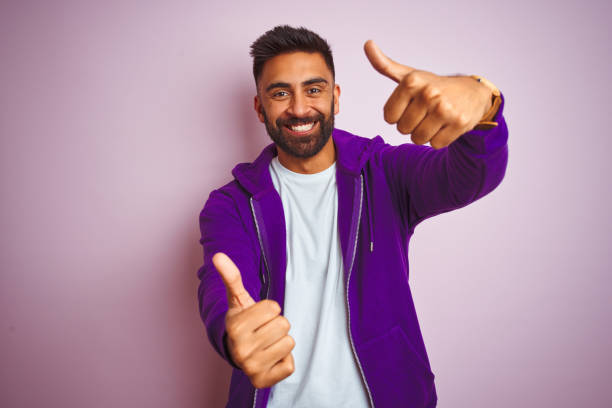 istock Young indian man wearing purple sweatshirt standing over isolated pink background approving doing positive gesture with hand, thumbs up smiling and happy for success. Winner gesture. 1167771034