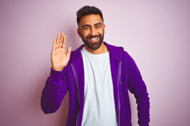 jeune homme indien utilisant le sweat-shirt pourpre restant au-dessus du fond rose d'isolement waiving disant bonjour heureux et souriant, geste amical de bienvenue - waving photos et images de collection