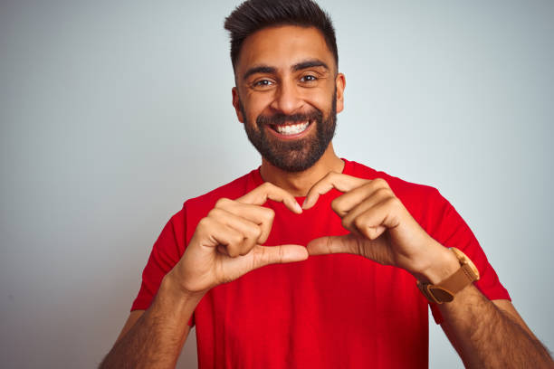 고립 된 흰색 배경 위에 빨간 티셔츠를 입은 젊은 인도 남자가 손으로 심장 기호와 모양을 보여주는 사랑에 미��소. 로맨틱 한 개념. - t shirt men red portrait 뉴스 사진 이미지