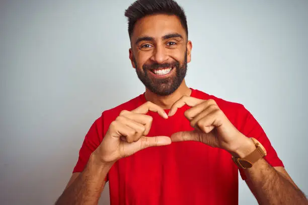 Photo of Young indian man wearing red t-shirt over isolated white background smiling in love showing heart symbol and shape with hands. Romantic concept.